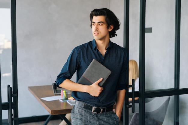 Medium shot portrait of serious businessman holding in hand stylish paper notebook standing in office thinking looking away Startup business team discussing project sitting at desk on background
