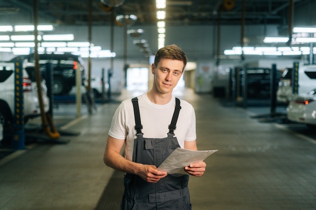 Medium shot portrait of smiling handsome young mechanic male wearing uniform holding clipboard stand...