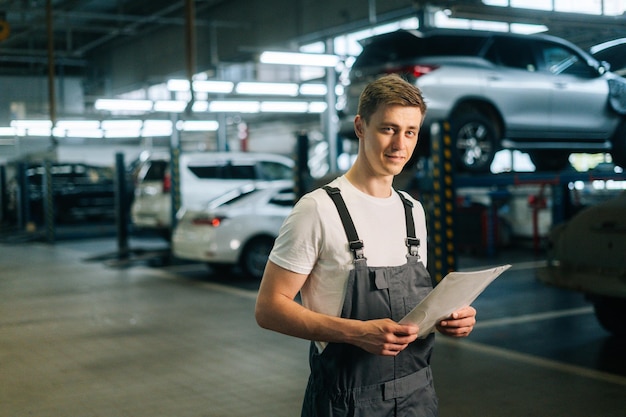 Ritratto medio di un bel giovane meccanico sorridente maschio che indossa l'uniforme che tiene appunti in piedi nel garage dell'officina riparazioni auto con sfondo del veicolo che guarda l'obbiettivo