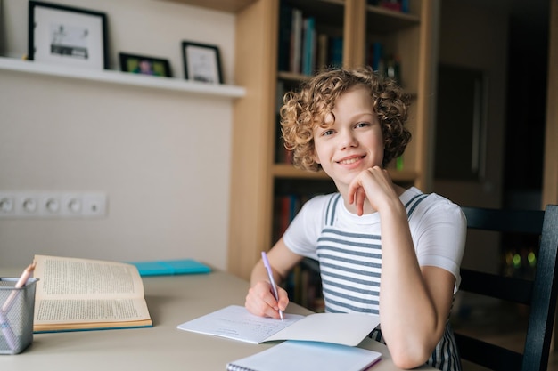 Medium shot portret van lachend krullend schoolmeisje dat thuis studeert en huiswerk maakt met pen en notities die naar de camera kijken