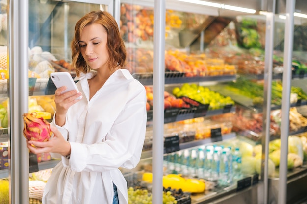 Medium shot van mooie jonge vrouw die exotisch fruit vasthoudt en op zoek is naar de naam op mobiele telefoon