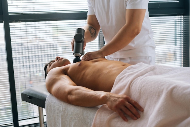 Medium shot van onherkenbare professionele mannelijke masseur die borstspieren ontvangt met behulp van massagepistool percussie-instrument van gespierde atleet man