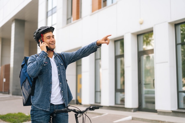 Medium shot van vrolijke knappe bezorger met thermische rugzak die op mobiele telefoon praat, ziet klant en markeert locatie door hand op te steken. Bezorger in beschermende helm die smartphone buitenshuis gebruikt.