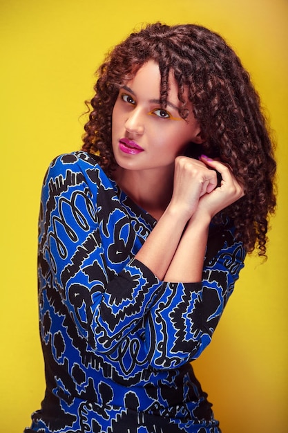 Medium shot of a woman with curly hair blue dress makeup and yellow background