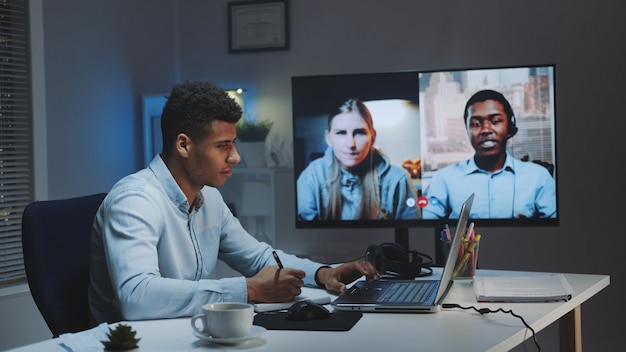 Medium shot of Young african chief manager making video call on big monitor with colleagues on quarantine