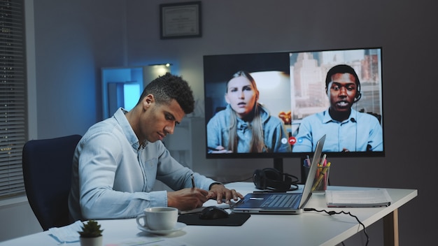 Photo medium shot of young african chief manager making video call on big monitor with colleagues on quarantine