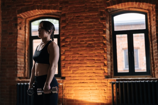 Medium shot of young athletic woman with beautiful strong body wearing activewear holding dumbbells during workout training. Caucasian fitness female workout out exercising in dark gym.