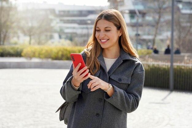 Medium shot of young smiling woman using cellphone in the street on sunny day