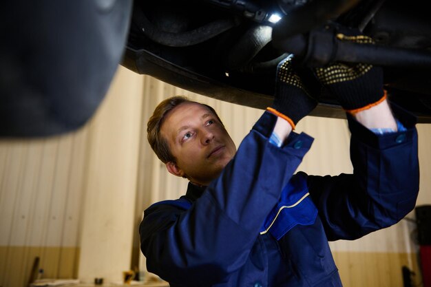 Meester automonteur in reparatieservice, technicus die de toestand onder de auto controleert, op voertuiglift in werkplaats. Monteur die de uitlijning van de auto controleert die het chassis controleert met behulp van een lamp.