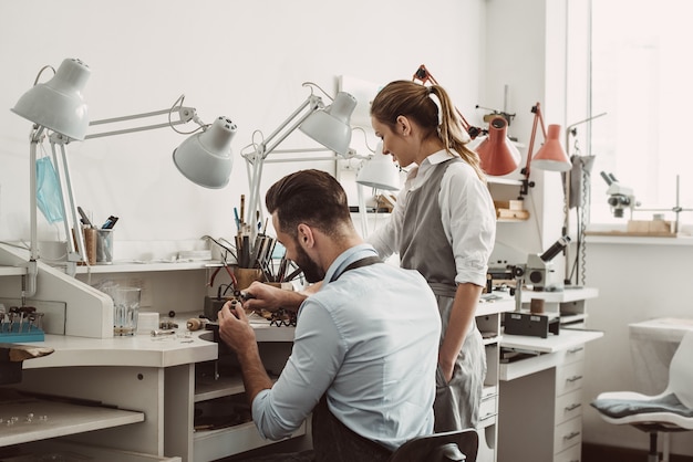 Meester en leerling. Jonge mannelijke assistent en vrouwelijke juwelier werken samen in een workshop sieraden maken. Bedrijf