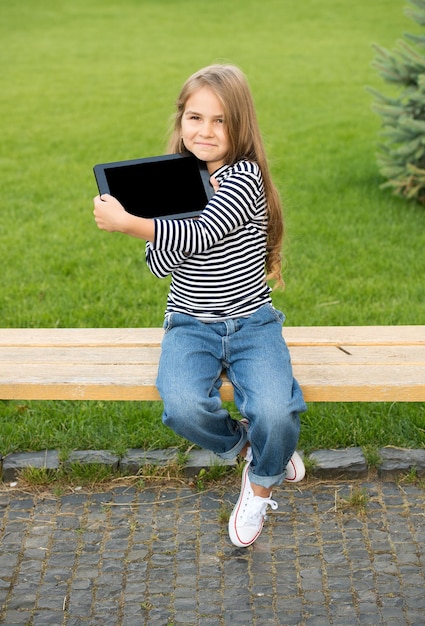 Познакомьтесь с ультрагладким дисплеем Счастливый ребенок показывает экран планшета, сидя на скамейке в парке Планшетный ПК Современный компьютер Сенсорная технология Мобильное устройство Интернет-серфинг Игра в онлайн-игры