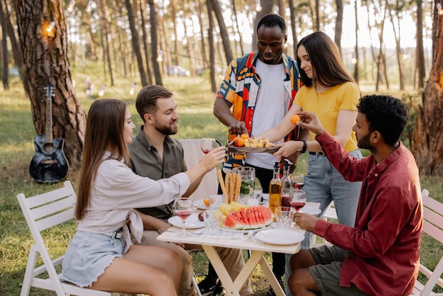 Meeting of multiracial group of friends eating dinner and drinking wine during party in the forest