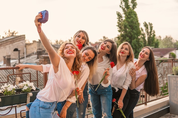 Meidenfeestje. Mooie vrouwenvrienden op het balkon of op het dak die selfie met bloemen nemen op vrijgezellenfeest. Ze dragen dezelfde kleren