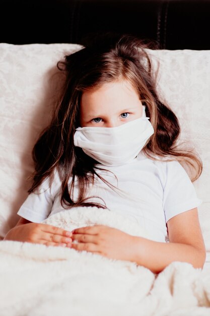 Meisje close-up zittend in een wit masker op het bed in de slaapkamer, zelfisolatie met een verkoudheid, de tweede golf van virussen.