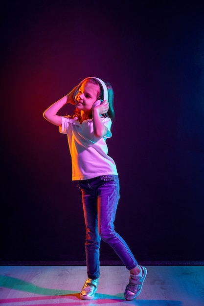 Meisje dat aan muziek in hoofdtelefoons op donkere kleurrijk luistert. Neonlicht. Dansend meisje. Gelukkig klein meisje dat op muziek danst. Leuk kind dat van gelukkige dansmuziek geniet.