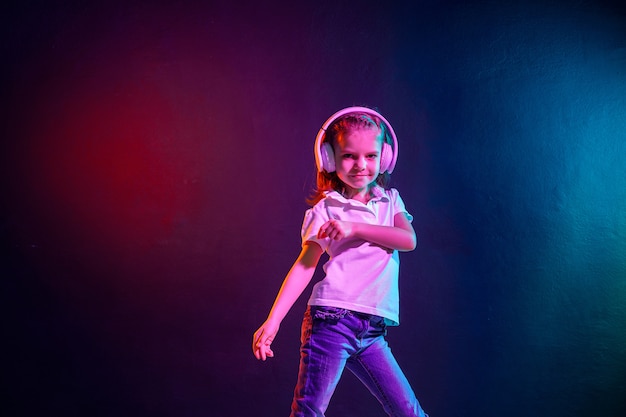 Meisje dat aan muziek in hoofdtelefoons op donkere kleurrijke muur luistert. Neonlicht. Dansend meisje. Gelukkig klein meisje dat op muziek danst. Leuk kind dat van gelukkige dansmuziek geniet.