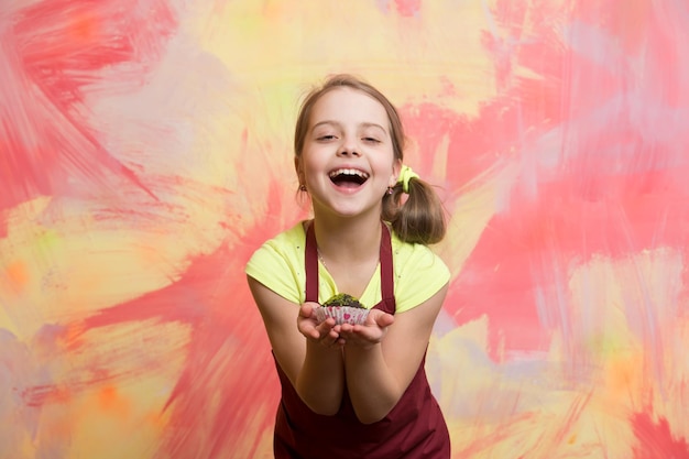 Meisje dat cupcake eet. Gelukkig kind lachend met taart. Babykok in rode chef-kokschort op kleurrijke abstracte muur. Bakken en heerlijke bakkerij. Gezond voedsel en dieetconcept.