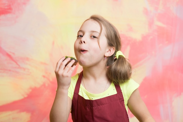 Meisje dat cupcake eet. Kind poseren met taart in mond. Bakken en heerlijke bakkerij. Babykok in rode chef-kokschort op kleurrijke abstracte muur. Gezond voedsel en dieetconcept.