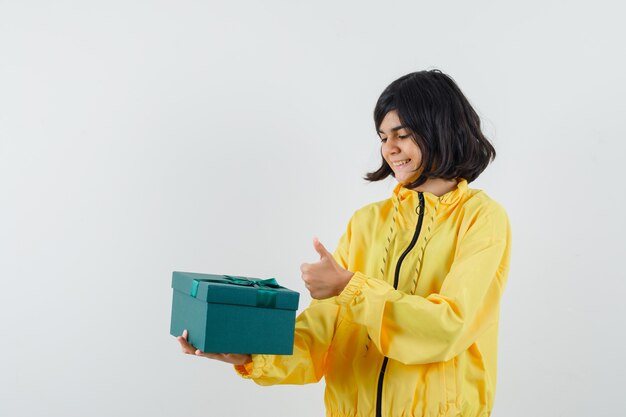 Meisje dat huidige doos houdt, duim in gele hoodie toont en vrolijk, vooraanzicht kijkt.