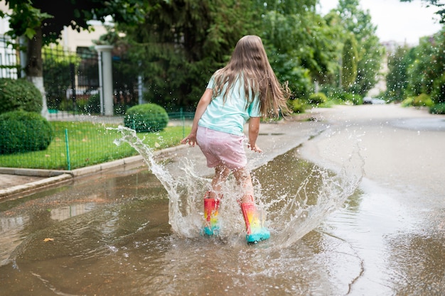 Meisje dat in regenboogrubberlaarzen in plassen dichtbij het huis springt