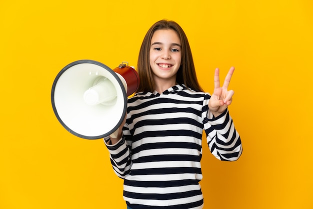 Meisje dat op gele achtergrond wordt geïsoleerd die een megafoon houdt en en overwinningsteken glimlacht toont