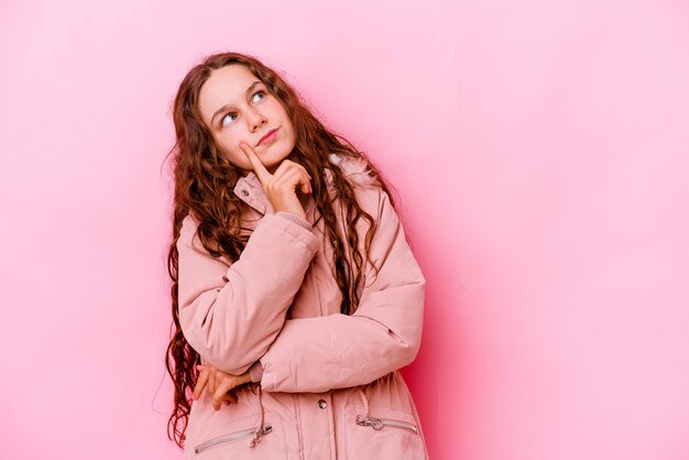 Meisje dat op roze muur wordt geïsoleerd die zijwaarts met twijfelachtige en sceptische uitdrukking kijkt