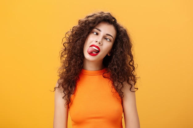 Meisje dat zorgeloos rondloopt en zich verveeld voelt terwijl ze zichzelf amuseert tijdens een saaie vergadering die tong uitsteekt, loensend en kantelend hoofd gedraagt zich kinderachtig en onvolwassen poseren over oranje achtergrond.