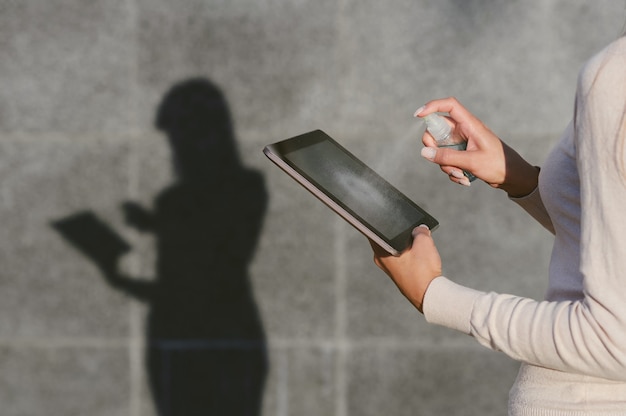 Meisje desinfecteert de tablet met antisepticum. Tegen de achtergrond van een grijze betonnen muur blijft een silhouet over met een duidelijke schaduw.