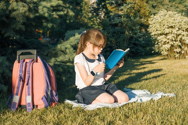 Meisje die een boek met een zitting van de schoolrugzak op het gras in het park lezen
