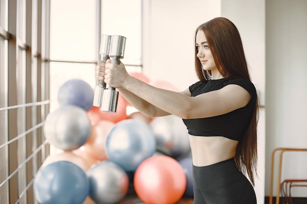 Meisje doet biceps met halters. Model met gemberhaar in zwarte sportkleding in een lichte sportschool. Fitnessballen trouwens.