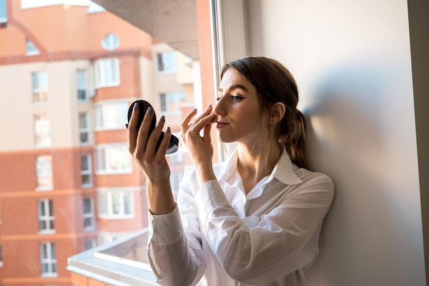 Meisje doet zelf make-up bij het raam op de achtergrond voordat ze een fotosessie neemt