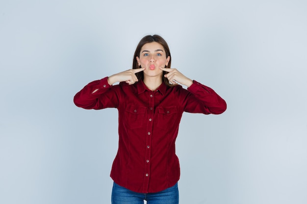 meisje drukt vingers op de wangen, pruilt lippen in een casual shirt en ziet er chagrijnig uit.