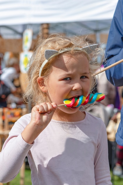 Meisje eet kleurrijk snoep. Een vijfjarig meisje in roze kleren eet een kleurrijk snoepje op een stokje. Zonnige zomerdag.