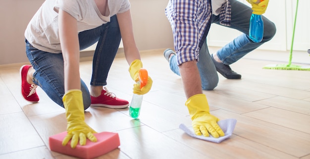 Meisje en jongen staan met één hand op de vloer. Ze dragen gele handschoenen. Het meisje houdt spons en bespuit met groene vloeistof. Man heeft blauwe doek en vloeistof. Weergave knippen.