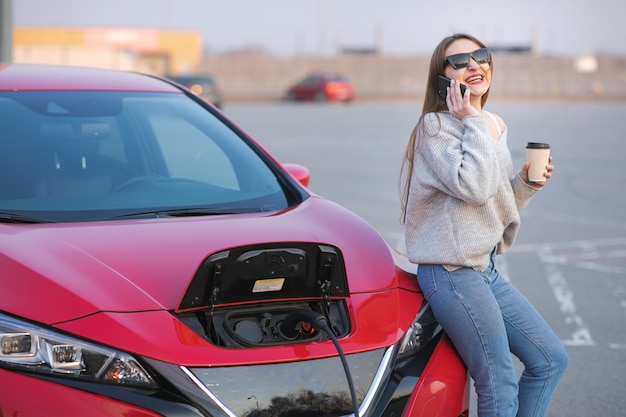 Meisje gebruikt koffiedrank tijdens het gebruik van een smartphone en wachtende voeding sluit aan op elektrische voertuigen om de batterij in de auto op te laden