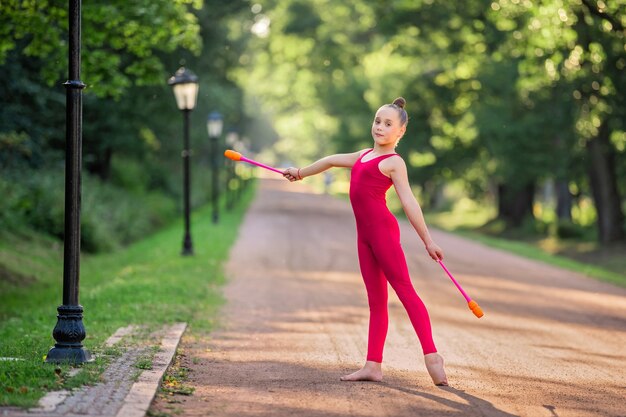 Meisje gymnast in heldere rode overall doet een oefening met clubs in het park op een warme zonnige avond