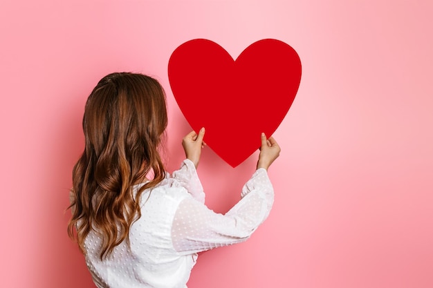 Meisje hangt een groot papieren hart aan een roze muur achteraanzicht Valentijnsdag concept