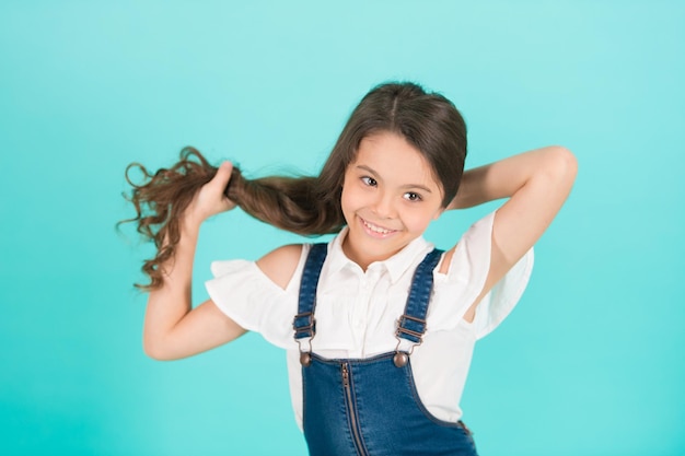 Meisje houdt lang haar op blauwe achtergrond Kindglimlach met gezond donkerbruin haar Haarverzorging kapselconcept Schoonheid of kapsalon punchy pastel