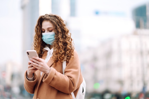 Meisje in beschermend steriel medisch masker op haar gezicht met een telefoon in quarantainestad. Vrouw die de telefoon gebruikt om naar nieuws te zoeken.