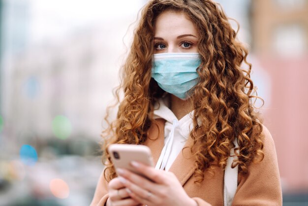 Meisje in beschermend steriel medisch masker op haar gezicht met een telefoon in quarantainestad. Vrouw die de telefoon gebruikt om naar nieuws te zoeken.