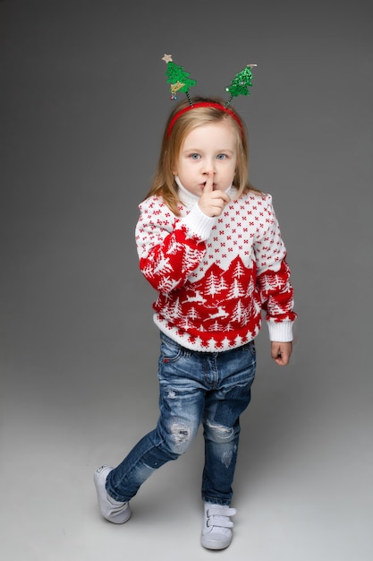 Meisje in blauwe gescheurde spijkerbroek en rode en witte trui die de vinger op haar lippen drukt.