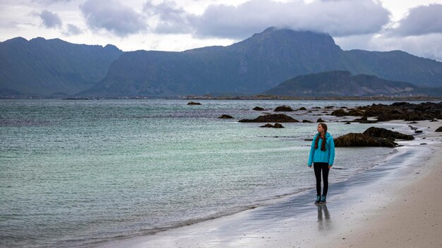 meisje in blauwe jas loopt op het strand met enorme rotsen en bergen op de achtergrond, Lofoten