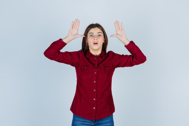 meisje in bordeauxrood shirt dat overgavegebaar toont en er bang uitziet.