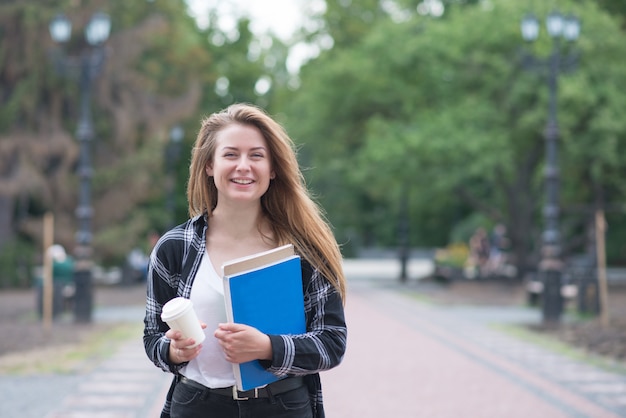 Meisje in casual kleding staat met boeken en notebooks op de achtergrond van het park, kijkt naar de camera