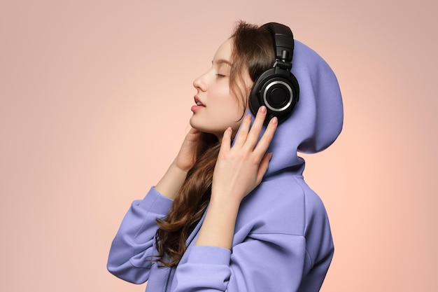 Meisje in draadloze koptelefoon, gezicht in profiel op een zachtroze achtergrond in een oversized hoodie.