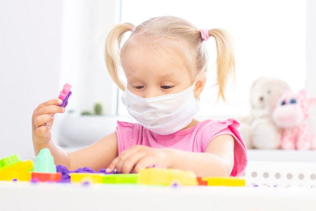 Meisje in een beschermend medisch masker speelt kinetisch zand in quarantaine. blond mooi meisje glimlacht en speelt met paars zand op een witte tafel. coronapandemie