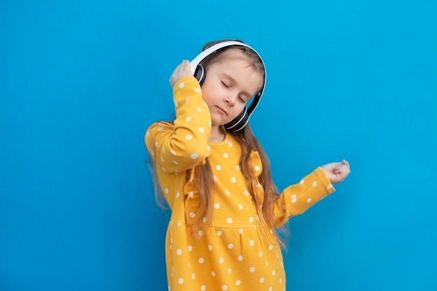 Meisje in een gele jurk luistert naar muziek in draadloze koptelefoons met haar ogen gesloten op een blauwe achtergrond Concept van kindervreugde