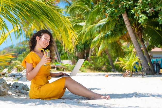 Meisje in een gele jurk op een tropisch zandstrand werkt op een laptop en drinkt verse mango. Werken op afstand, succesvolle freelance. Werkt op vakantie.