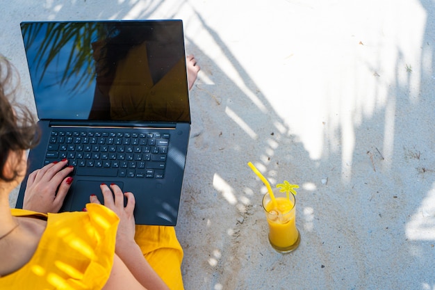 Meisje in een gele jurk op een tropisch zandstrand werkt op een laptop en drinkt verse mango. Werken op afstand, succesvolle freelance. Werkt op vakantie.