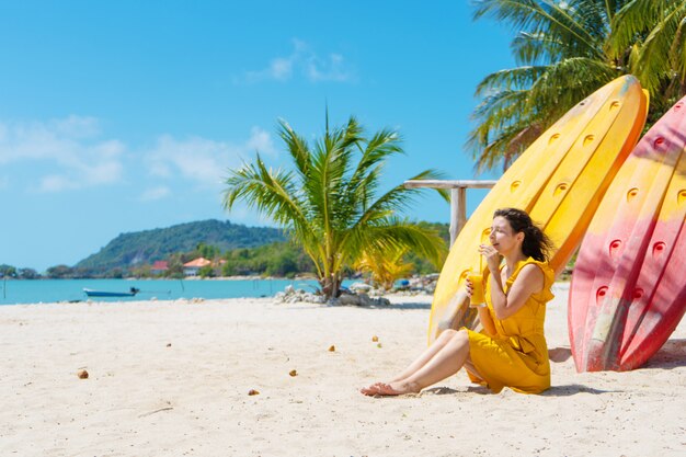 Meisje in een gele jurk op een tropisch zandstrand werkt op een laptop in de buurt van kajaks en drinkt verse mango. Werken op afstand, succesvolle freelance. Werkt op vakantie.